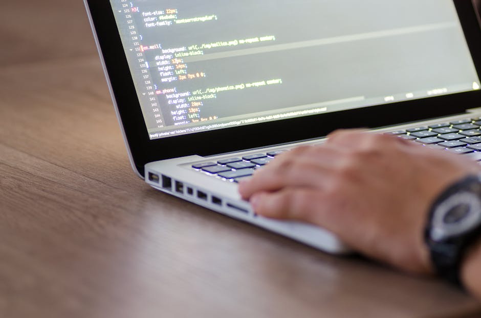 A close-up view of a person's hand resting on a laptop keyboard displaying lines of code with highlighted syntax, placed on a wooden desk. The individual is wearing a black wristwatch with a round dia