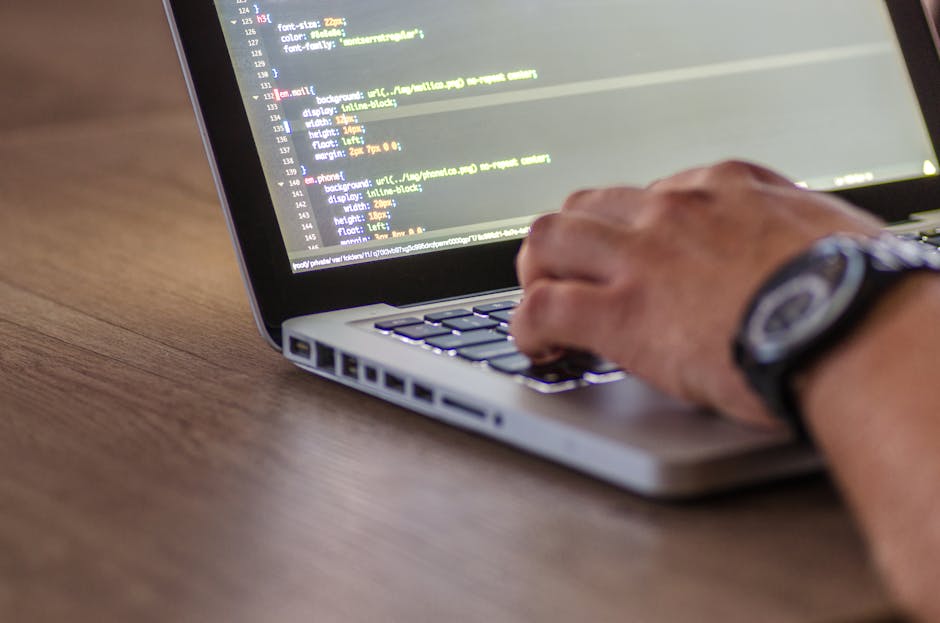 A close-up view of a person's hand resting on a laptop keyboard displaying lines of code with highlighted syntax, placed on a wooden desk. The individual is wearing a black wristwatch with a round dia