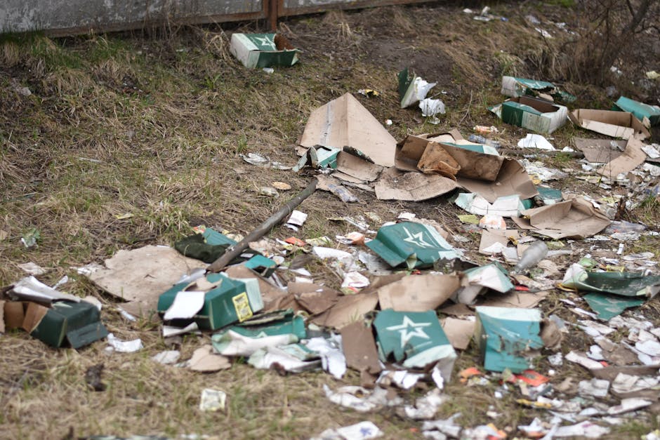A patch of a garden or outdoor area with patchy grass and exposed soil, littered with scattered cardboard boxes, some flattened and torn, along with pieces of paper and plastic debris. The background 