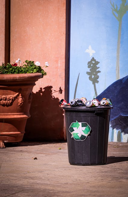A patch of a garden or outdoor area with patchy grass and exposed soil, littered with scattered cardboard boxes, some flattened and torn, along with pieces of paper and plastic debris. The background 
